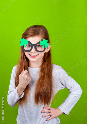 Portrait of little smiling caucasian red-haired girl in mask of leprechaun shamrock clover glasses for irish St. Patrick's Day on green studio background. Copyspace.