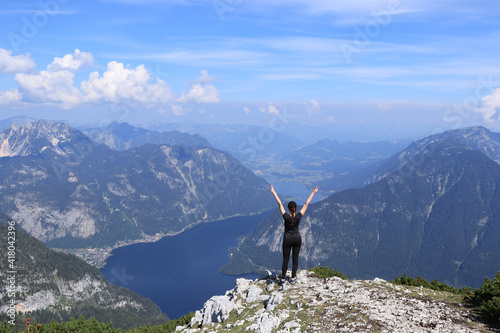 girl in black sportswear rejoices in defeating the Dachstein Krippenstein and enjoys the view of Hallstatter See and the adjacent village of Hallstatt with her hands over her head. © Fauren