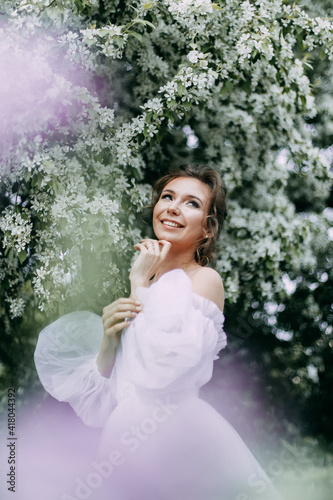 A beautiful gentle attractive young woman in a wedding dress with a bouquet of lilacs walks alone and poses in a spring garden with flowering plants