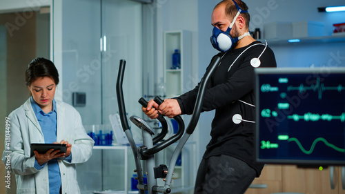 Monitor showing EKG reading of athlete running on cross trainer, while medical specialists supervise exercise, controlling physical activity, measuring heart rate in sports science laboratory photo