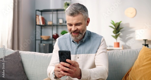 Close up of cheerful handsome middle-aged Caucasian man in positive mood texting on smartphone while sitting at modern home on sofa and smiling, grey-haired male using social network app on cellphone photo