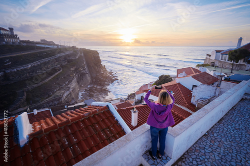 Traveling by Portugal. Young woman taking photo of wonderful sunset on rock ocean shore.