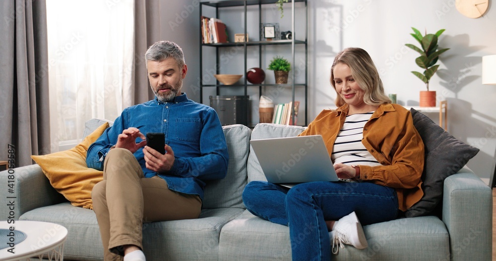 Obraz premium Happy married couple resting at home on sofa in living room using digital gadgets. Middle-aged Caucasian man texting on smartphone while his pretty wife typing on laptop browsing online, tech concept