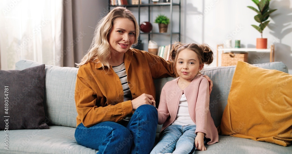 Portrait of Caucasian happy positive beautiful mother and cute little daughter girl sitting at home on couch in cozy living room looking at camera and smiling, family relations, parenthood concept