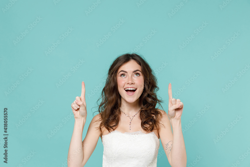 Excited bride young woman 20s in beautiful white wedding dress pointing index fingers overhead up isolated on blue turquoise color wall background studio portrait. Ceremony celebration party concept. Excited bride young woman 20s in beautiful white wedding dress pointing index fingers overhead up isolated on blue turquoise color wall background studio portrait. Ceremony celebration party concept.
