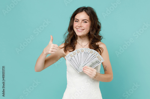 Smiling bride young woman in white wedding dress hold fan of cash money in dollar banknotes showing thumb up isolated on blue turquoise background studio portrait. Ceremony celebration party concept.