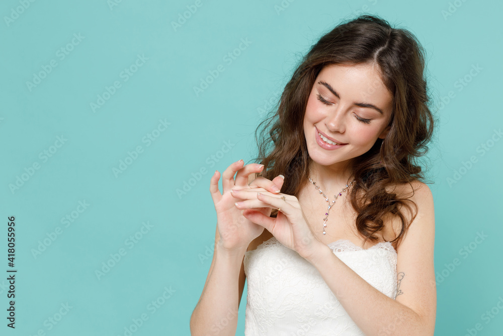 Smiling charming bride young woman 20s in beautiful white wedding dress looking at hand finger with ring isolated on blue turquoise background studio portrait. Ceremony celebration party concept. Smiling charming bride young woman 20s in beautiful white wedding dress looking at hand finger with ring isolated on blue turquoise background studio portrait. Ceremony celebration party concept.