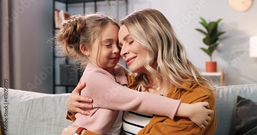 Close up of cute nice loving Caucasian little girl child hugging mother in living room at home. Caring mom embracing small daughter of sofa in apartment, love and care, family time concept