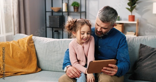 Portrait of joyful cute Caucasian little daughter and caring father spending time together at home videochatting on tablet online. Dad and small child girl watching cartoons on device, gadget digital