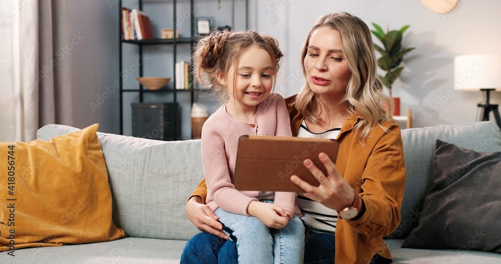 Portrait of happy joyful caring Caucasian mom with small daughter in room together videochatting using tablet. Pretty mother teaching small girl to use gadget device, family time, watching cartoons Portrait of happy joyful caring Caucasian mom with small daughter in room together videochatting using tablet. Pretty mother teaching small girl to use gadget device, family time, watching cartoons