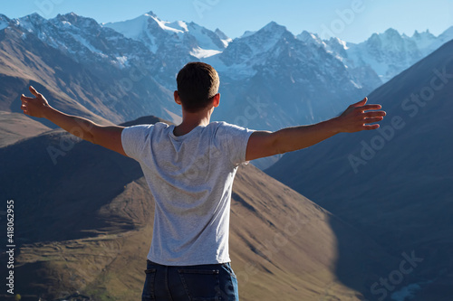 Valokuva Athletic guy in white t-shirt stands on hilltop and looks at large mountains pea