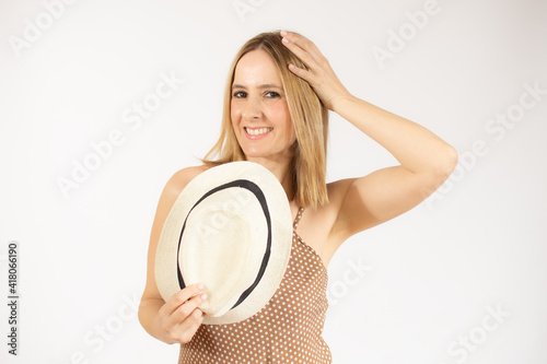 Pleased elegant woman in dress and straw hat posing while looking at camera over white background