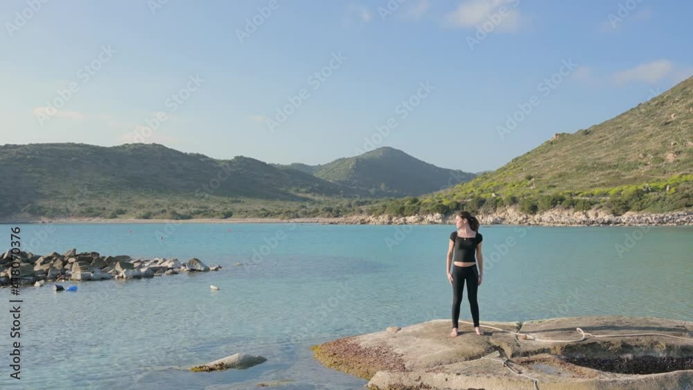 Young woman in suit jump practicing sport in nature, standing on a rock with beautiful sea background with translucent water. Relaxing with cervical spine stretches. Young woman in suit jump practicing sport in nature, standing on a rock with beautiful sea background with translucent water. Relaxing with cervical spine stretches.