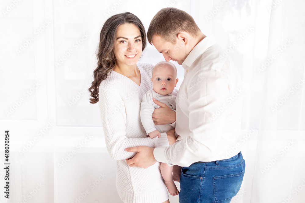 family, the concept of a happy young family with a small child, father, mother and daughter hugging at home by the window family, the concept of a happy young family with a small child, father, mother and daughter hugging at home by the window