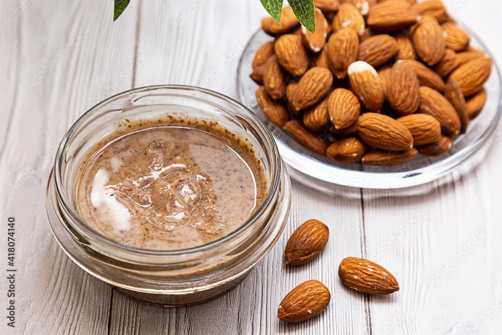 A jar of almond butter with a bunch of fresh almond on a white wooden table with an olive branch. Homemade aimond butter, natural, healthy food. The modern wellness and vegan concept. A jar of almond butter with a bunch of fresh almond on a white wooden table with an olive branch. Homemade aimond butter, natural, healthy food. The modern wellness and vegan concept.