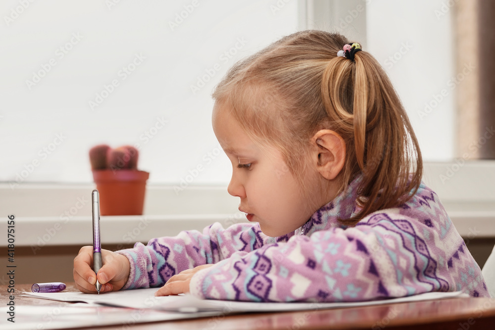Little girl doing homework. The child sits at the table and writes with a pen in a notebook. Home schooling for a little girl Little girl doing homework. The child sits at the table and writes with a pen in a notebook. Home schooling for a little girl