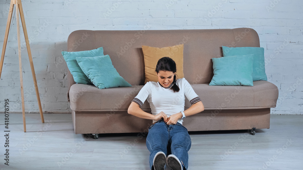 Fototapeta premium young woman sitting and fastening jeans in living room