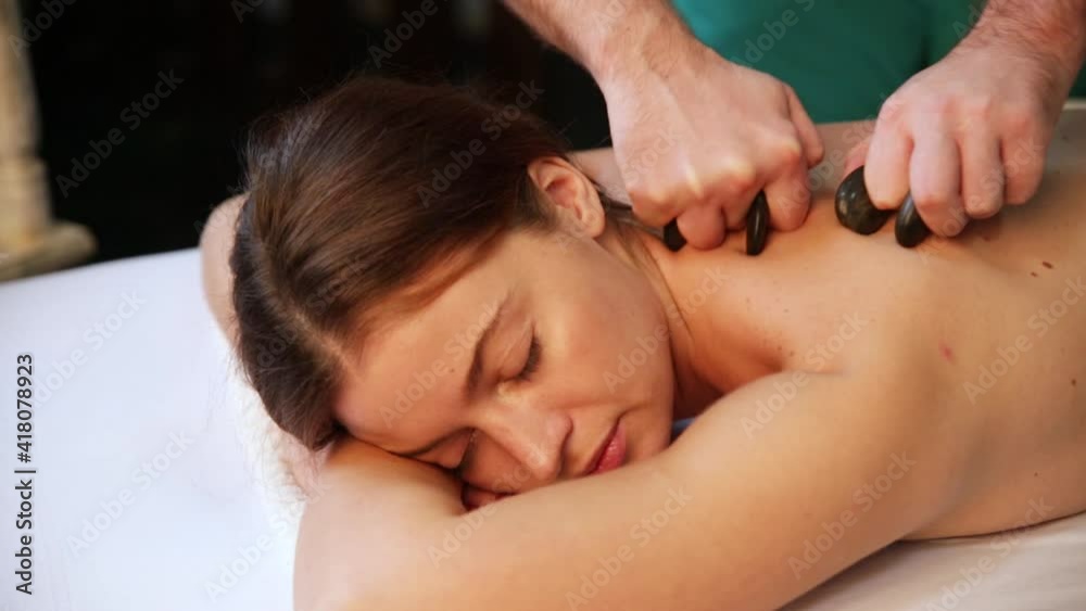 A beautiful girl lies on her stomach during a stone massage. Hands of a massage therapist massaging the shoulder. A beautiful girl lies on her stomach during a stone massage. Hands of a massage therapist massaging the shoulder.