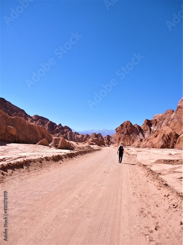 Red dirt road in the desert on Chile with rocky mountains on both sides, a man walking on it and blue sky. 