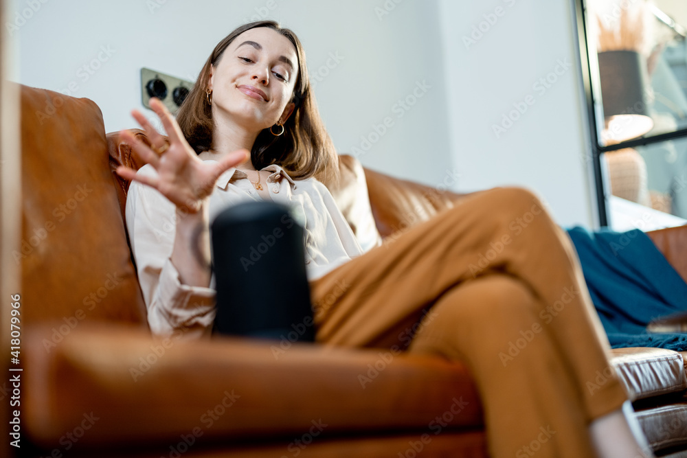 Happy pretty woman speaking with black audio assistant column on the coffee table while sitting on brown leather sofa in stylish living room. Smart home concept. Happy pretty woman speaking with black audio assistant column on the coffee table while sitting on brown leather sofa in stylish living room. Smart home concept.