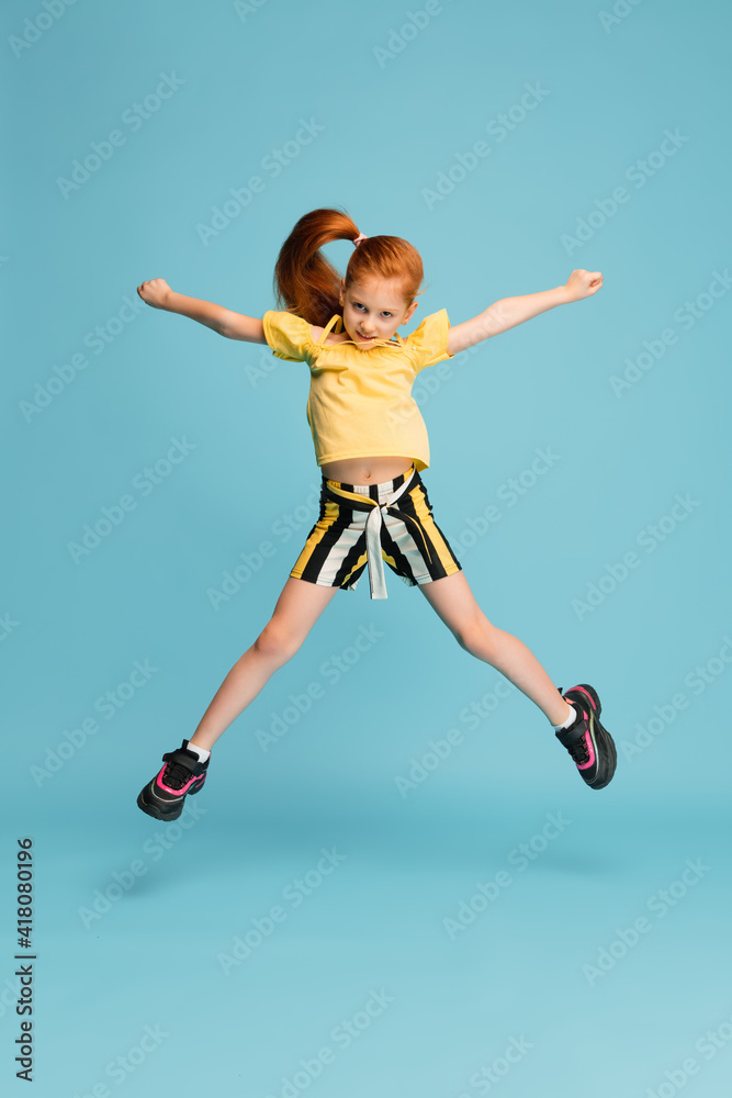 Jumping high. Happy, smiley little caucasian girl isolated on blue studio background with copyspace for ad. Looks happy, cheerful. Childhood, education, human emotions, facial expression concept.