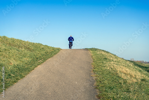 Unidentified man cycles on the driveway to the road on top of the Dutch dike. The photo was taken on a sunny day in the winter season with a clear blue sky.