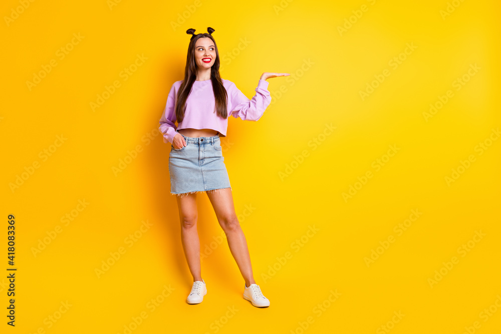 Full size photo of optimistic pretty brunette girl look hold empty space wear pink sweater skirt sneakers isolated on yellow background