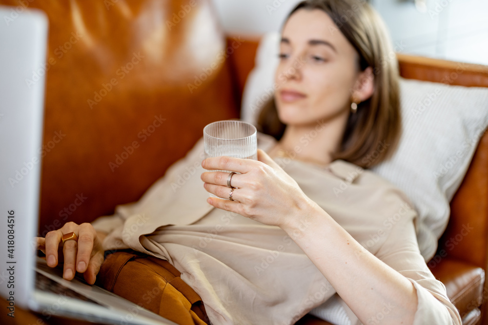 Pretty woman hold a glass of water while working on laptop and lying on sofa. Lifestyle and healthy concept. Dehydration. Rate of drinking water per day. Focus on the glass. Pretty woman hold a glass of water while working on laptop and lying on sofa. Lifestyle and healthy concept. Dehydration. Rate of drinking water per day. Focus on the glass.