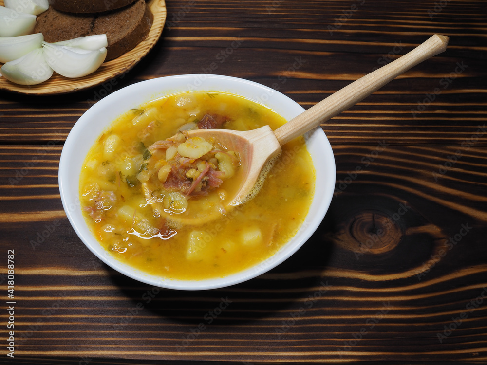 Pea soup with vegetables in a white bowl, wooden spoon, plate with rye bread and onions in a wooden plate on a wooden table, top view. Healthy hot pease dish for lunch Pea soup with vegetables in a white bowl, wooden spoon, plate with rye bread and onions in a wooden plate on a wooden table, top view. Healthy hot pease dish for lunch