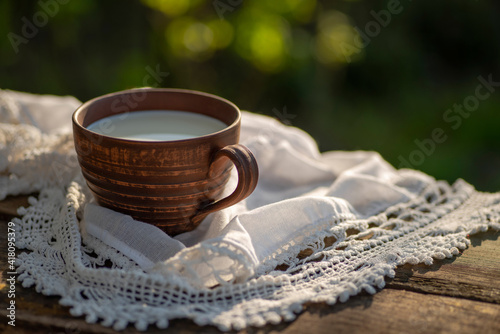 Pottery cup with milk (kefir, yogurt, sour cream, kumis), lace tablecloth, wooden table. Outdoor picnic, breakfast, brunch, refreshments. Soft focus
