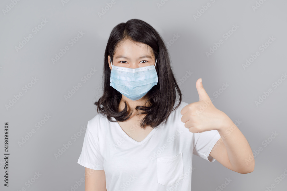 Portrait of Asian teenager in medical face mask to protect Covid-19 (Coronavirus) wears white t-shirt showing thumb up on gray background, people to stay home, health and medicine concept Portrait of Asian teenager in medical face mask to protect Covid-19 (Coronavirus) wears white t-shirt showing thumb up on gray background, people to stay home, health and medicine concept