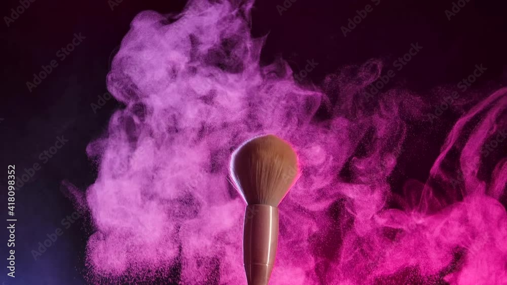 Two makeup brushes collide and cause a swirl of powder particles against a black background in pink neon light. Fine dust of the crushed powder flies beautifully in the air. Close up. Slow motion.
