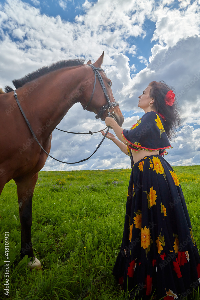 Obraz premium Beautiful gypsy girl with a horse in field with green glass in summer day and blue sky and white clouds background. Model in ethnic dress posing with farm animal