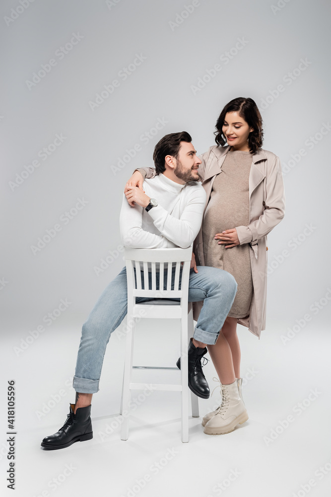 Smiling pregnant woman holding hand of husband on chair on grey background Smiling pregnant woman holding hand of husband on chair on grey background
