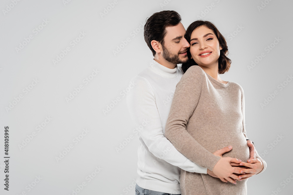 Fototapeta premium Happy man hugging cheerful pregnant woman isolated on grey