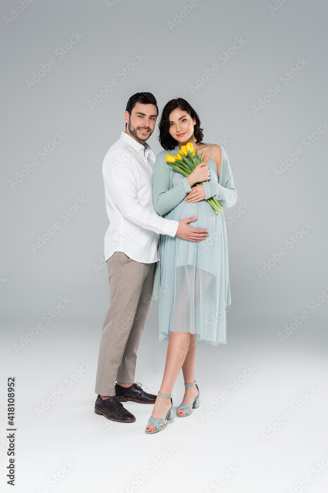 Cheerful man hugging pregnant wife in dress holding flowers on grey background