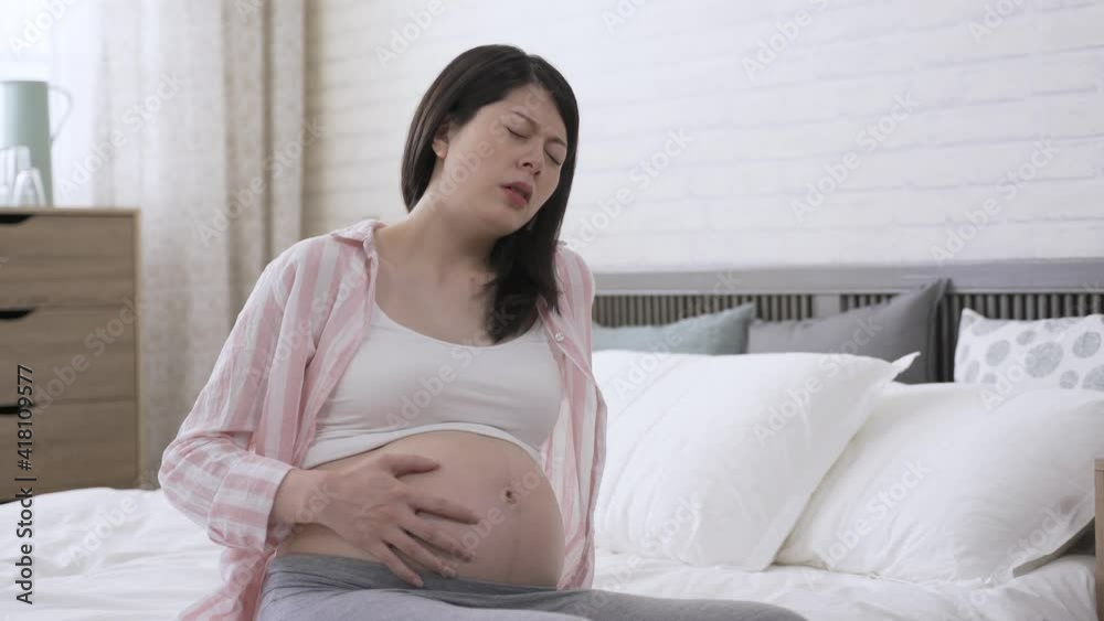 pregnant female is leaning backward to support herself with hand on bed while feeling strong contraction and groaning in pain at home. pregnant female is leaning backward to support herself with hand on bed while feeling strong contraction and groaning in pain at home.