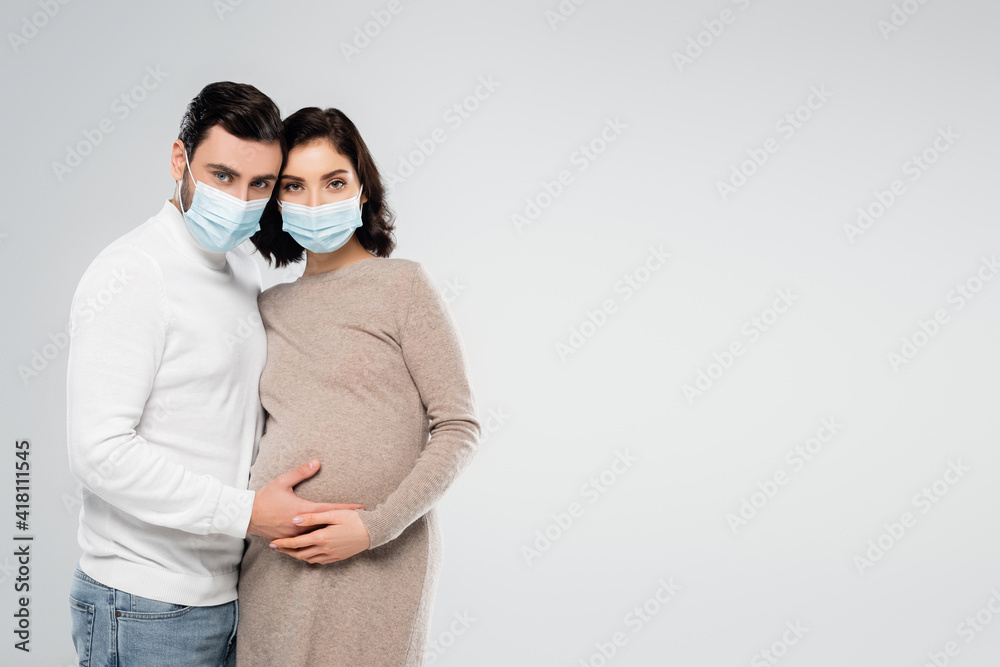 Man in medical mask touching belly of pregnant wife isolated on grey