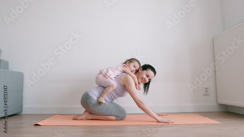 Baby girl sits on her mother's back who exercises on yoga mat at home