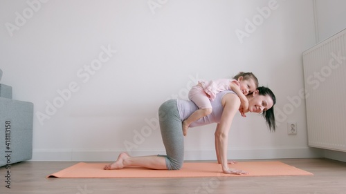 Baby girl fun and sits on her mother's back who exercises on yoga mat at home