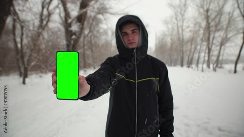 Man in park winter shows screen of phone of chomakey. Man holds a phone with a green screen in hand the nature of winter. photo