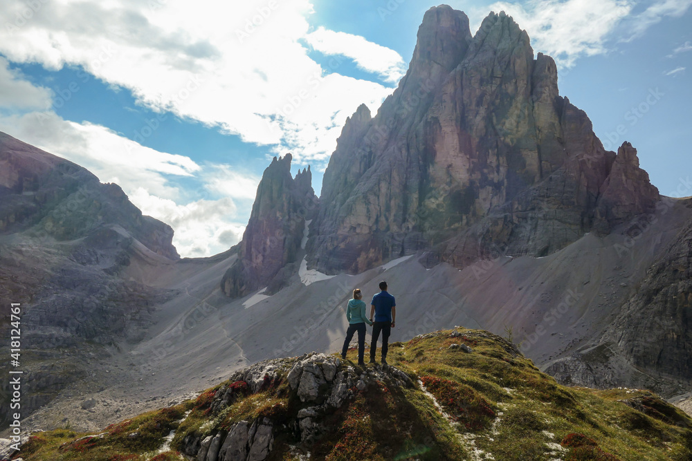 Couple standing under the foot of high Italian Dolomites and enjoying the panoramic. There are many sharp peaks in front. Lots of lose stones and landslides. Sharing experience. Happiness and love