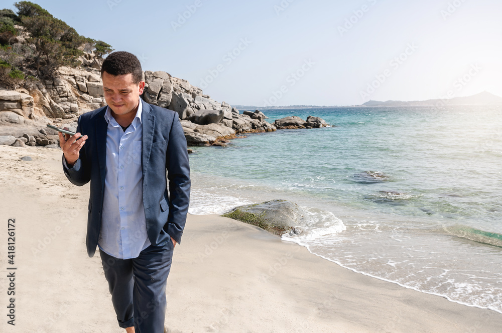Latin businessman with suit walking on beautiful sandy beach while holding a mobile phone and listening a vocal message Latin businessman with suit walking on beautiful sandy beach while holding a mobile phone and listening a vocal message