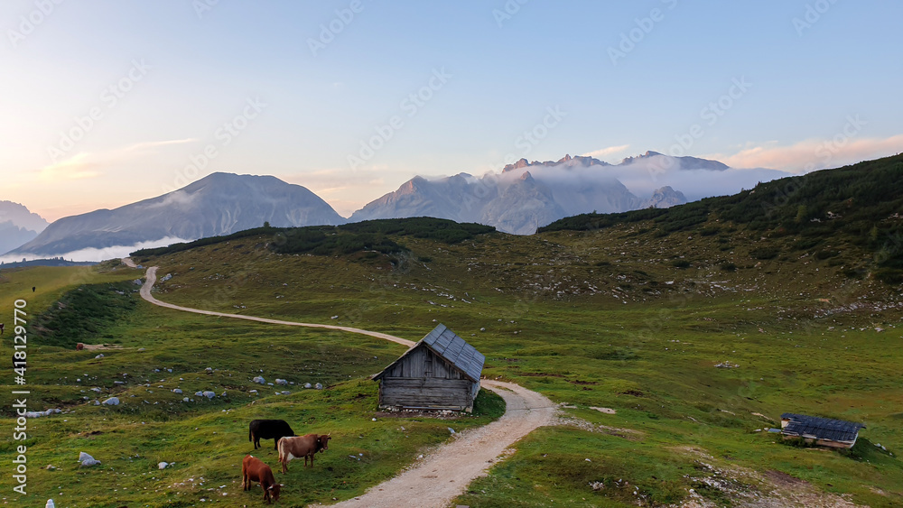 An early morning in Italian Dolomites. There is a small wooden cottage on a green meadow. In the back there are high mountain chains, bursting with sunrise colors. New day beginning. Golden hour.
