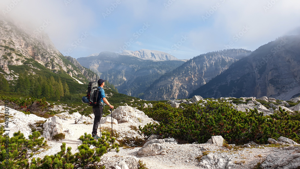 A man with a big hiking backpack enjoying the panoramic view on Italian Dolomites. He stands on a stony plateau, above the valley. A few clouds around. In the back high mountain chains. Freedom