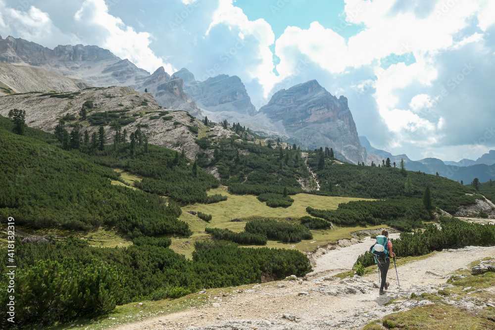 A woman with a hiking backpack hiking on a gravelled road in high Italian Dolomites. There are a few trees on the lower parts, and steep and sharp mountain chain in the back. Discovering and exploring