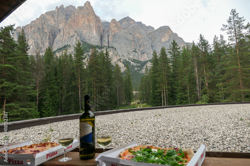 A bottle of Italian wine and two pizzas next to it placed on a small balcony with the view on high Italian Dolomites. There is a dense forest at the bottom of the valley. Cloudy day. Italian cuisine photo