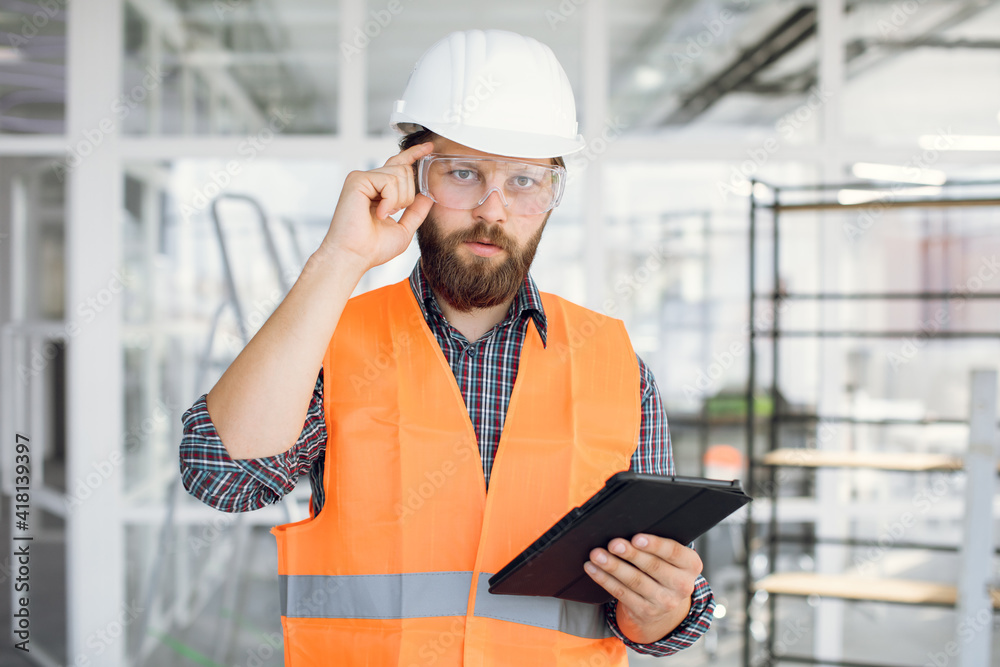 Competent arhitect posing indoors with digital tablet in hands. Bearded man wearing uniform, protective glasses and construction hardhat. Competent arhitect posing indoors with digital tablet in hands. Bearded man wearing uniform, protective glasses and construction hardhat.