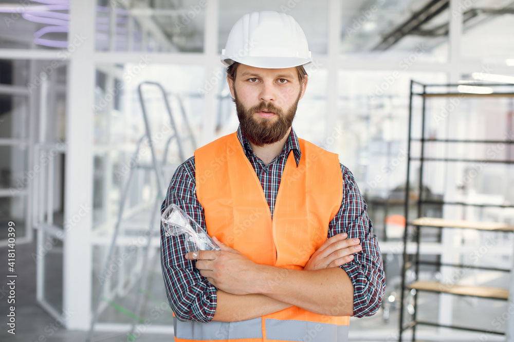 Portrait of bearded man wearing uniform and white hardhat at work. Confident contractor standing with crossed arms on building object.