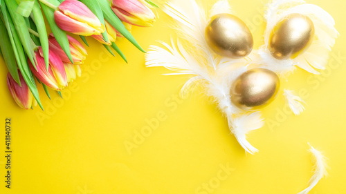 Basket easter decoration: Golden eggs in basket with spring tulips, white feathers on pastel yellow background. Congratulatory easter design. Top view.